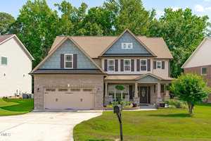 This charming two-story house at 1498 Fireside Lane in Creedmoor boasts a brick and blue-toned exterior, complete with a two-car garage, driveway, and cozy front porch adorned with seating and potted plants. Surrounded by a well-manicured lawn, it's nestled among trees and neighboring homes.