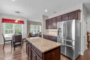 Modern kitchen with dark wood cabinets, a stainless steel refrigerator, and a large granite island. A cozy dining area with a round table and chairs by a window is in the background. Warm lighting creates an inviting atmosphere.