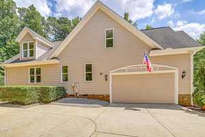 A beige two-story house with a gable roof features a two-car garage adorned with an American flag. The driveway is curved, with shrubbery on the side. Surrounded by trees, the house sits under a partly cloudy sky.