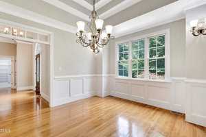 A bright, empty dining room with hardwood floors, wainscoting, and a tray ceiling. Large windows let in natural light, illuminating the elegant chandelier. A doorway leads to another room.