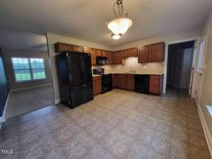 A spacious kitchen featuring dark wood cabinets, black appliances, and a tiled backsplash. A modern light fixture hangs from the ceiling, illuminating the tile floor. An open doorway leads to a carpeted living area with a large window.