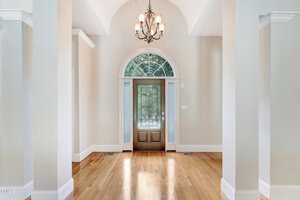 Spacious and elegant entryway featuring a wooden door with glass panels and an arched window above. The foyer has light-colored walls, a high ceiling, and a classic chandelier, with polished hardwood floors adding warmth to the space.