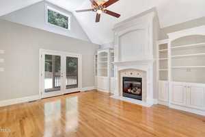 A spacious living room features a vaulted ceiling with a fan, a large white fireplace flanked by built-in shelves, and hardwood floors. A set of French doors leads to an outdoor deck, and natural light illuminates the space.