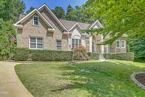 A large brick and stucco house with a gabled roof sits proudly at 2020 Silverleaf Drive, surrounded by manicured hedges and trees. A driveway leads to the front entrance, where an American flag waves near the door. The setting in Youngsville is bright under a clear blue sky.
