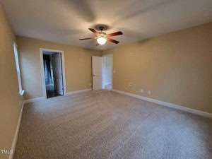 A spacious empty room with beige walls and carpeted floor. A ceiling fan with lights is centered on the ceiling. An open white door leads to another room with a patterned curtain visible. Two windows allow natural light in.