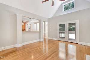 Spacious, bright room with polished hardwood floors and high, vaulted ceiling. Features a ceiling fan, large windows with a sliding glass door, and a view into a kitchen through an open partition. Walls painted light gray.