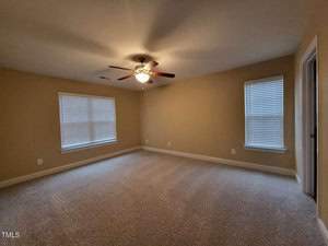 A carpeted room with beige walls and two windows with blinds. A ceiling fan with a light is centered on the ceiling. There are two doorways visible, one open on the right side of the room. The space appears empty and well-lit.