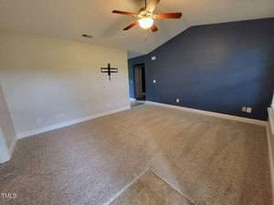 A spacious empty living room with beige carpet, a ceiling fan, and a dark blue accent wall. The room has white walls, a cross decoration, tiled flooring near an entrance, and ample natural light from an unseen window.