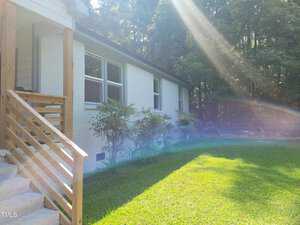 A white brick house with large windows framed by wooden railings on the front steps. Sunlight streams across the green lawn, creating a rainbow effect. Tall trees are in the background.