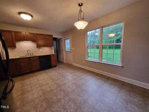 A kitchen with brown cabinets, black appliances, and a beige tile backsplash. The floor is tiled, and large windows overlook a grassy yard. Two ceiling lights illuminate the room, and a door leads outside.