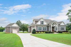 A large suburban house with beige siding, white trim, and a gray roof, surrounded by a well-manicured lawn. There's a separate garage to the left and a long driveway lined with greenery under a partly cloudy sky.