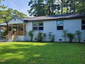 A white brick house at 308 9th Street, featuring a black shingled roof, is surrounded by green grass and shrubs. It has multiple windows and a covered porch with wooden railings. Tall trees rise in the background under a clear blue sky, embodying the charm of Butner.