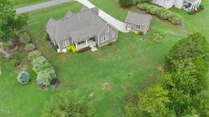 Aerial view of a suburban house with a brown roof, surrounded by a large green lawn, trees, and shrubs. The driveway leads to a garage. Nearby, there are additional houses and well-maintained landscaping.