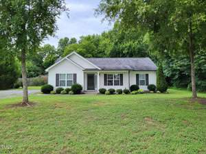 Nestled at 2211 Irvin Street, this small, single-story white house with a gray roof is surrounded by lush greenery and bushes. Set on a neatly manicured lawn with trees in the background under a cloudy sky, it embodies the charm of Oxford's serene landscapes.