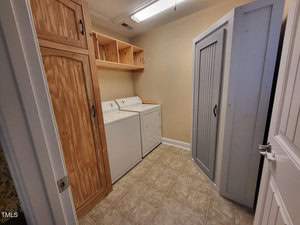 A laundry room with beige walls features a washer and dryer side by side. Wooden cabinets and shelves are mounted above the appliances, and a gray utility cabinet stands to the right. The floor is covered with beige tiles.