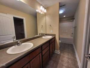 A bathroom with dual sinks on a long countertop, wooden cabinets, and a large mirror. The floor is tiled, and in the background, there's a bathtub with a showerhead and a closed closet door. The walls are painted beige.