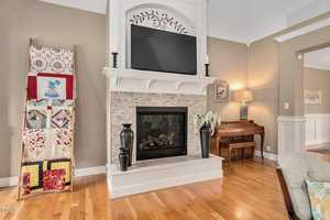A cozy living room with a stone fireplace and a TV mounted above it. A quilt ladder displays colorful quilts to the left, while a wooden piano sits in the corner. A lamp and floral arrangement add warm touches to the room's decor.