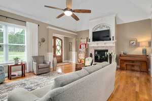 A cozy living room with a gray sectional sofa facing a fireplace and TV. The room features a ceiling fan, a piano, and a large window with curtains. A patterned rug, an armchair, and decorative plants are present. A wooden door is in the background.