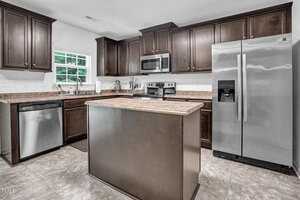Modern kitchen with dark wood cabinets, stainless steel appliances, a central island with a brown countertop, and tile flooring. There is a window over the sink, and the room has a clean and organized feel.