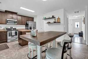 A modern kitchen with dark wood cabinets, stainless steel appliances, and a rustic wooden table with high chairs. The room is bright, with open shelves displaying dishes and decor. A hallway leads to a white front door.