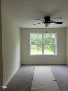 A bright, unfurnished room with light-colored walls and carpeted flooring. A large window with tree views lets in natural light. A ceiling fan is centered above, and a sheet of paper is laid out on the floor.