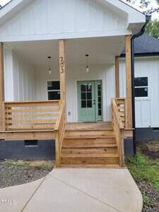 Wooden porch with stairs leads to a mint green door of a white house in Butner. The porch features two hanging lights and a horizontal railing, while "703 W D Street" is prominently displayed on a wooden post. The sidewalk gracefully curves towards the entrance.