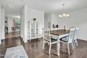 A dining room with white walls and hardwood floors features a white dining table with six chairs. A decorative chandelier hangs above the table. A glass cabinet and small side table are in the background, with natural light from a window.