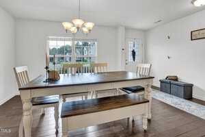 A dining room with a wooden table and matching bench, surrounded by chairs. A chandelier hangs above the table. The room features a large window, hardwood floor, decorative wall sign, and a cushioned storage bench near the door.