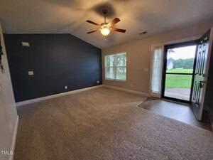 A carpeted room with a ceiling fan, featuring neutral walls and a dark accent wall. Large windows provide ample natural light. An open door leads outside to a grassy area, visible through the glass screen door.