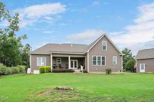 A single-story house with a gray exterior and a small front porch is set in a grassy yard. The sky is partly cloudy. A garage is visible on the right, with an American flag displayed. Trees are in the background.