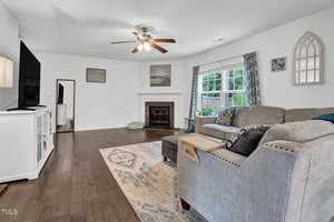 A cozy living room with a large gray sectional sofa, a wooden coffee table, and a patterned rug. A flat-screen TV is mounted on a white stand. The room features a fireplace, wall art, and two windows with blue curtains, letting in natural light.