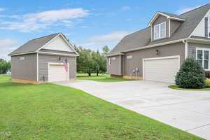 A suburban home with gray siding, a large driveway, and a neatly trimmed lawn. An American flag is mounted on a smaller adjacent shed. The sky is clear and blue, providing a calm atmosphere.