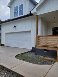 Modern two-story house with white siding and a black roof. Features a two-car garage and a wooden porch with railing. The driveway and walkway are paved, with a small patch of greenery near the entrance.