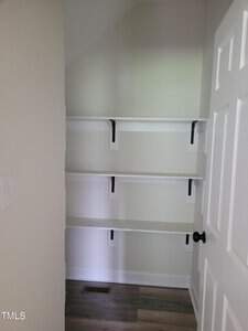 A small, empty closet with three white shelves supported by black brackets. The walls are light-colored, and the floor is wooden. A white door with a black round handle is partially open, revealing the closet's interior.