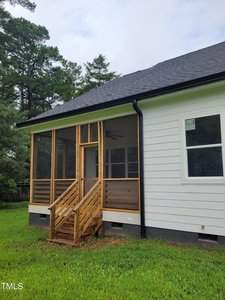 A white house with a black roof featuring a wooden screened porch and steps leading to a grassy yard. Surrounding the house are tall evergreen trees under a cloudy sky.