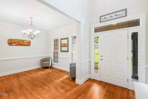 A bright entryway with hardwood floors opening into a living room. A chandelier hangs above two gray armchairs near a window. A "Bless This Home" sign is above the white front door, which overlooks a green lawn.