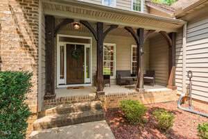A welcoming front porch with wooden beams and a brick exterior featuring a dark wooden door adorned with a wreath. Two wicker chairs with cushions sit on the porch, surrounded by shrubs and red mulch landscaping.