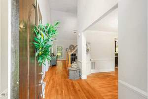 A bright, welcoming hallway features wooden floors and a high ceiling. A staircase with a wooden banister is visible on the right. A green wreath decorates the open front door, and light streams in from a window in the background.