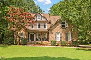 A large two-story brick house on 3819 St Lucy Drive, Franklinton, boasts a charming porch and multiple windows, nestled amid lush green grass and trees. To the left of the entrance blooms a striking flowering tree with pink blossoms.