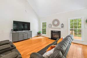 A bright living room with a vaulted ceiling, featuring a large flat-screen TV on a gray console. There's a fireplace surrounded by windows, a dark sofa with colorful pillows, and wooden floors. The room opens to an outdoor deck.