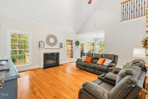 A spacious living room with high ceilings and wooden floors. It features a fireplace with a round mirror above, large windows with natural light, and two dark leather sofas with orange cushions. Stairs lead to an upper level, and greenery is visible outside.