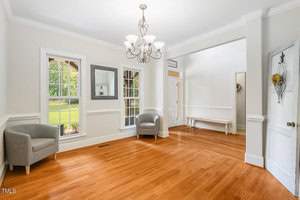 A bright room with wooden floors, two gray chairs by large windows, and a white bench in the corner. A decorative chandelier hangs from the ceiling. A white door with a floral arrangement is partially open, revealing a hallway.