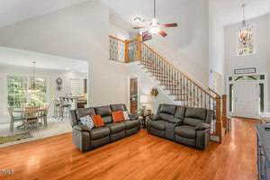 A spacious, open-concept living room with wooden floors, two black leather sofas, and a staircase leading to an upper level. Large windows let in natural light, and an adjoining dining area is visible. A ceiling fan hangs from the high ceiling.