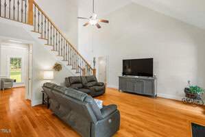 A spacious living room with a high ceiling and wooden floors features a large TV on a console, two black leather sofas, and a ceiling fan. A staircase with a wooden railing and iron balusters leads upstairs. Natural light enters through a doorway.