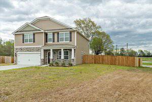 Two-story house with beige siding and stone accents. It features a two-car garage and a covered front porch. A wooden fence encloses the backyard. Cloudy sky and trees in the background.