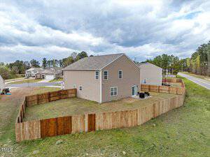 A two-story beige house with a gray roof, enclosed by a wooden fence. The property has a grassy yard and is situated near a road and other homes, surrounded by trees under a cloudy sky.