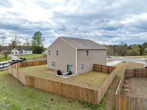 A two-story beige house with a fenced backyard, set in a suburban neighborhood. The backyard features patio furniture on a grassy lawn. The sky is cloudy, and other houses and trees are visible in the background.