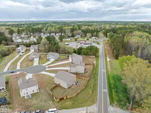 Aerial view of a suburban neighborhood with several houses. A cul-de-sac is on the left, with homes surrounding it. A road runs along the right side, bordered by trees, and there are fenced backyards. The sky is cloudy.