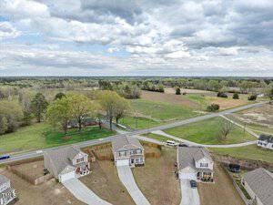 An aerial view of a suburban neighborhood with a row of similar houses, each with driveways, surrounded by green fields and trees. A road divides the residential area from open farmland under a partly cloudy sky.