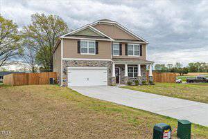 A two-story house with a brown and stone exterior, featuring a two-car garage and a large front lawn. The sky is overcast, and there's a wooden fence surrounding the yard. A mailbox is visible at the edge of the driveway.